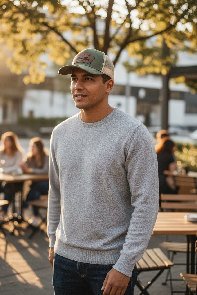 Man wearing a cap and gray sweater standing outdoors with people and tables in the background
